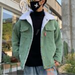 Homme portant une veste en velours verte et un tee-shirt noir, il est dans une rue.