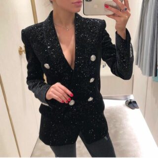 blazer en velours noir brillant pour femme porté par une femme prenant un selfie avec son téléphone portable devant un miroir, dans une pièce ressemblant à une chambre d'hôtel avec des murs couleurs crème et une moquette grise au sol.