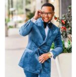 homme noir souriant, portant un ensemble en velours bleu contenant un blazer et un pantalon, des lunettes carrées et un noeud papillon, dans une rue sur un trottoir spacieux, à côté d'une plante