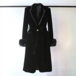 On voit un manteau long noir, en velours, suspendu à une tringle noire. Les manches sont décorées de fausses fourrures et le col en V est très élégant.