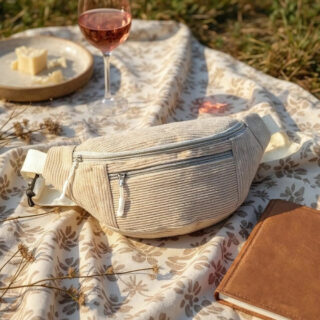 Sac banae en velours beige à deux fermetures zippées posée sur un tapis de pique-nique avec un livre marron, un assiette et un verre de vin.