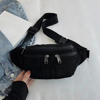 Sac banane en velours trois poches zippées de couleur noire posée sur in tissu blanc avec une veste en jean à gauche et une main.
