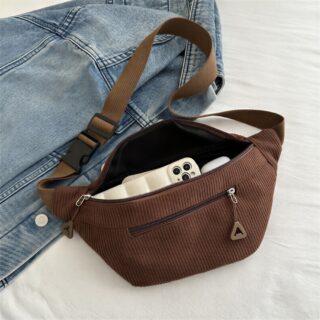 Un sac banane bandoulière en velours côtelé femme marron, posé sur un jean bleu, avec une poche zippée et une sangle réglable.
